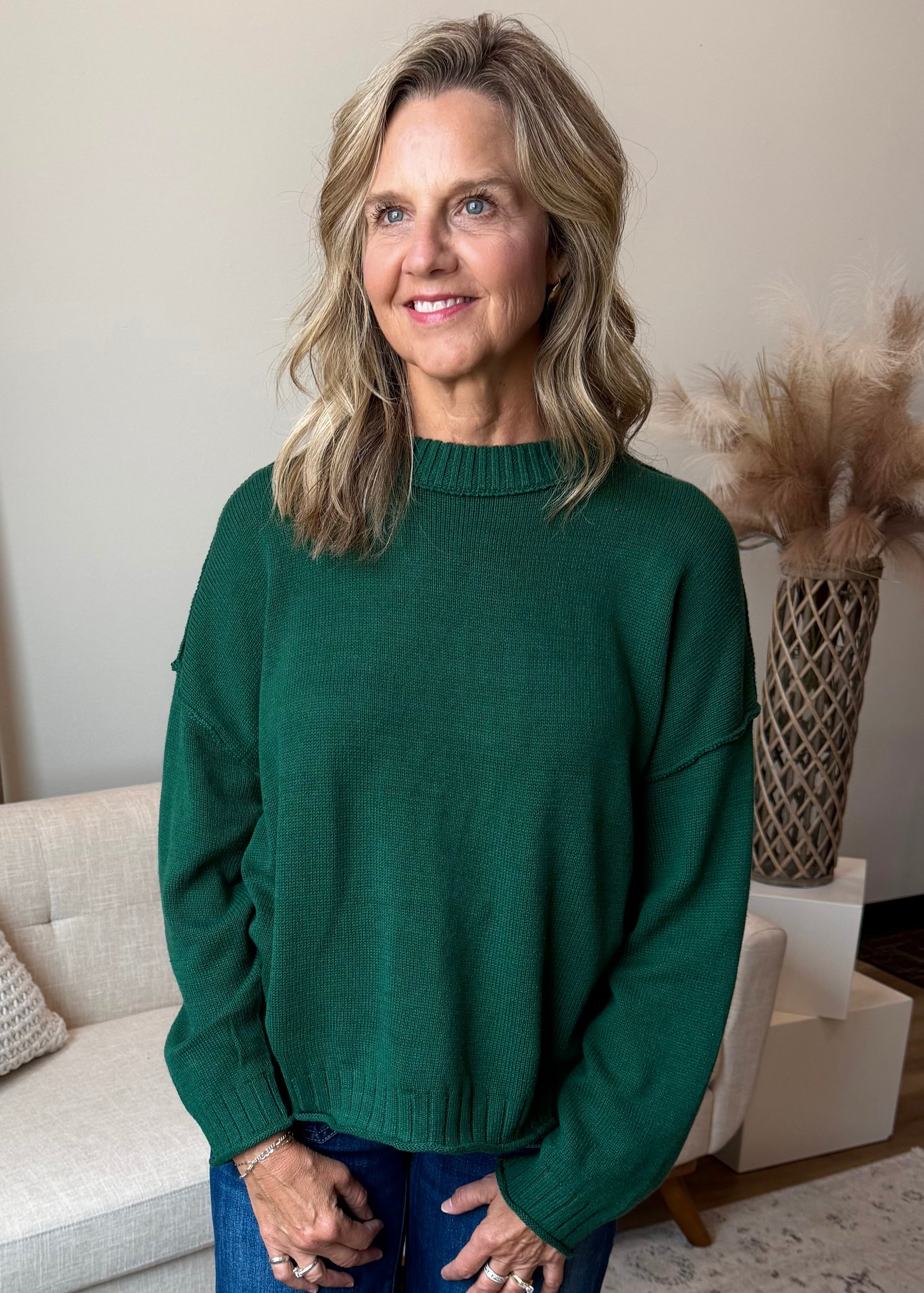 A woman stands in a styled living room wearing a dark green knit sweater and blue jeans, with a white sofa, wooden ladder, and patterned rug in the background.