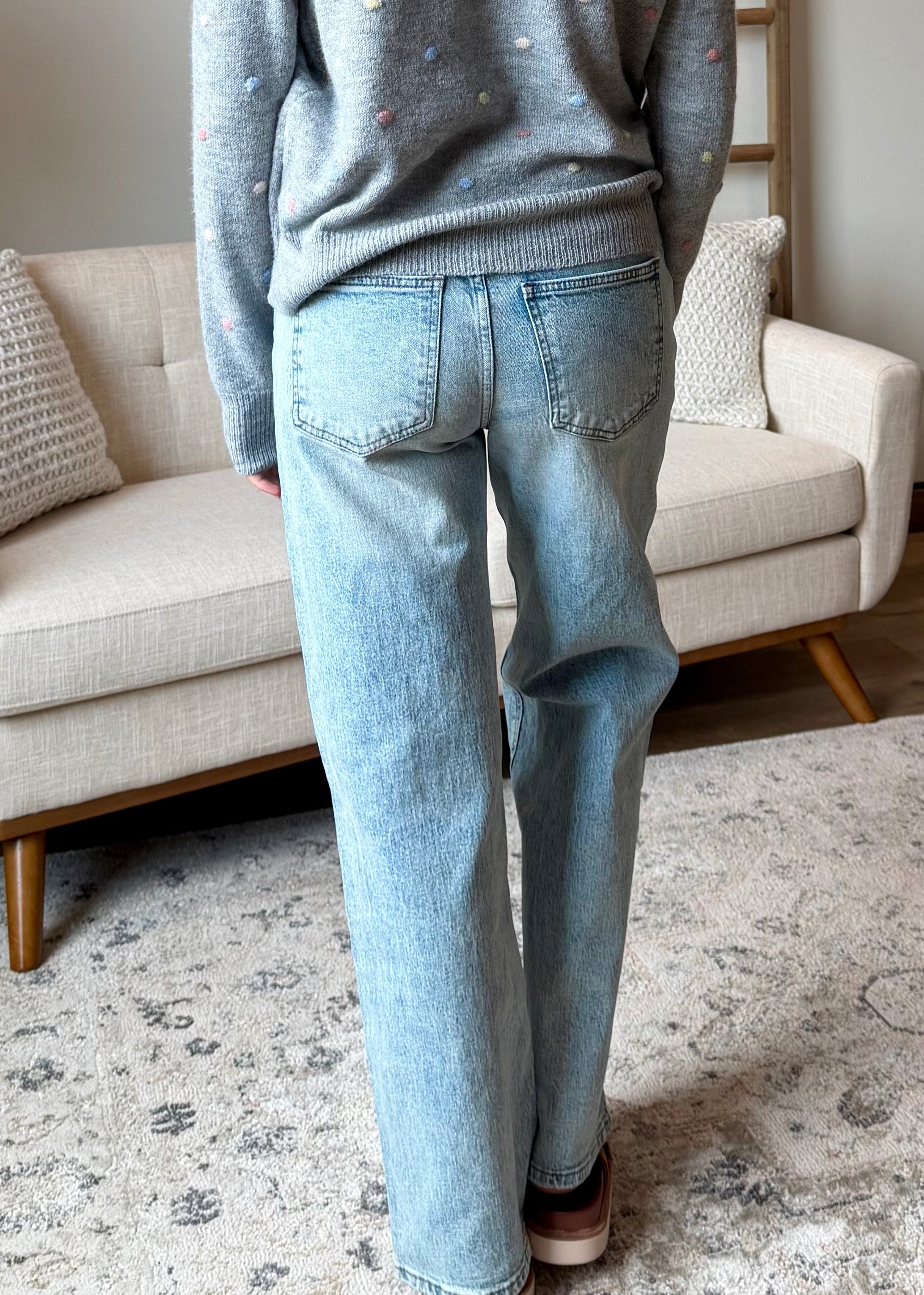 A woman stands with her back to the camera in a bright, modern living room, wearing a light gray knit sweater with subtle white speckles and light-wash blue jeans.
