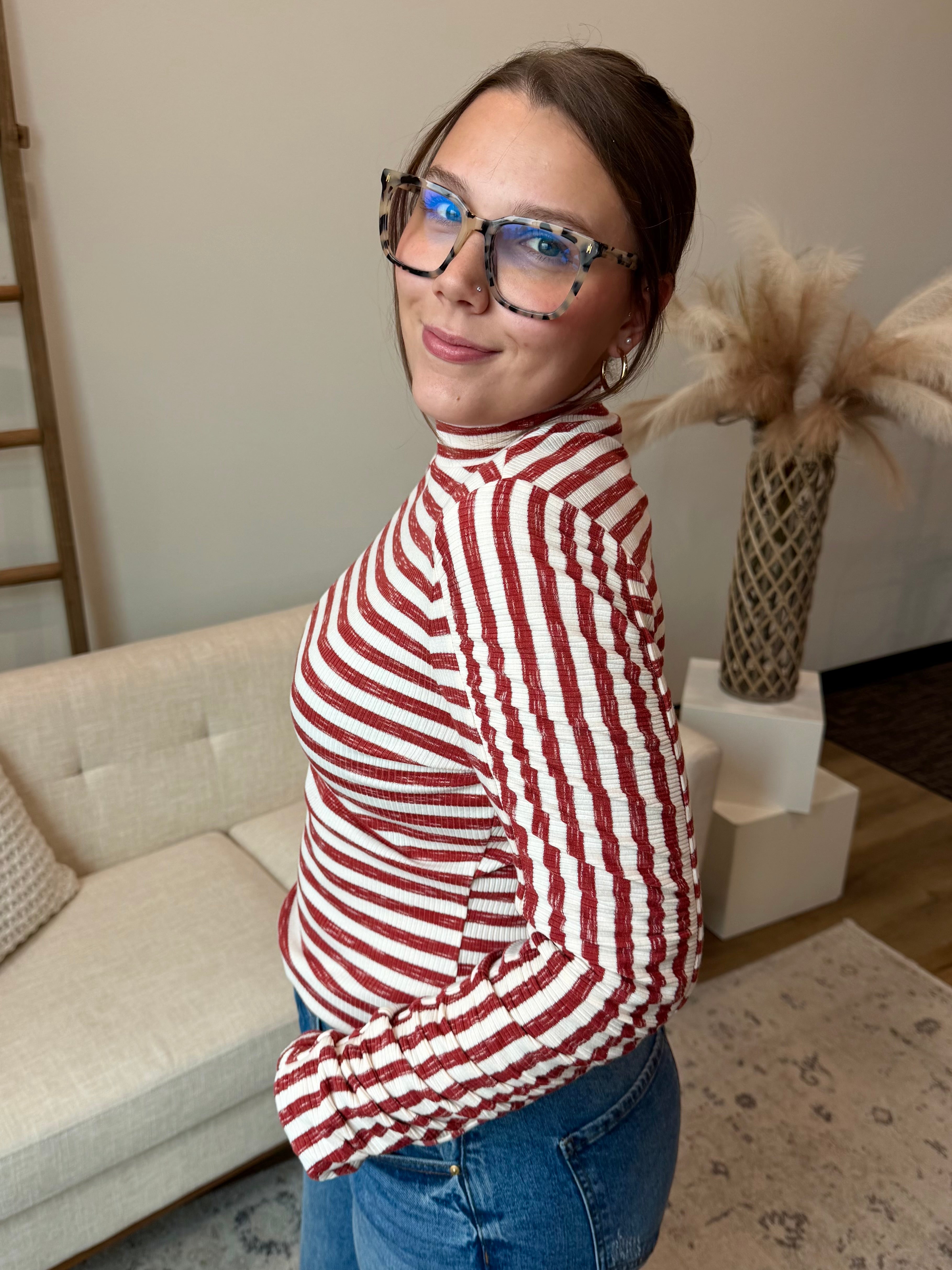 A woman wearing a red and white striped long-sleeve mock neck top and blue jeans, standing in a modern living room setting.