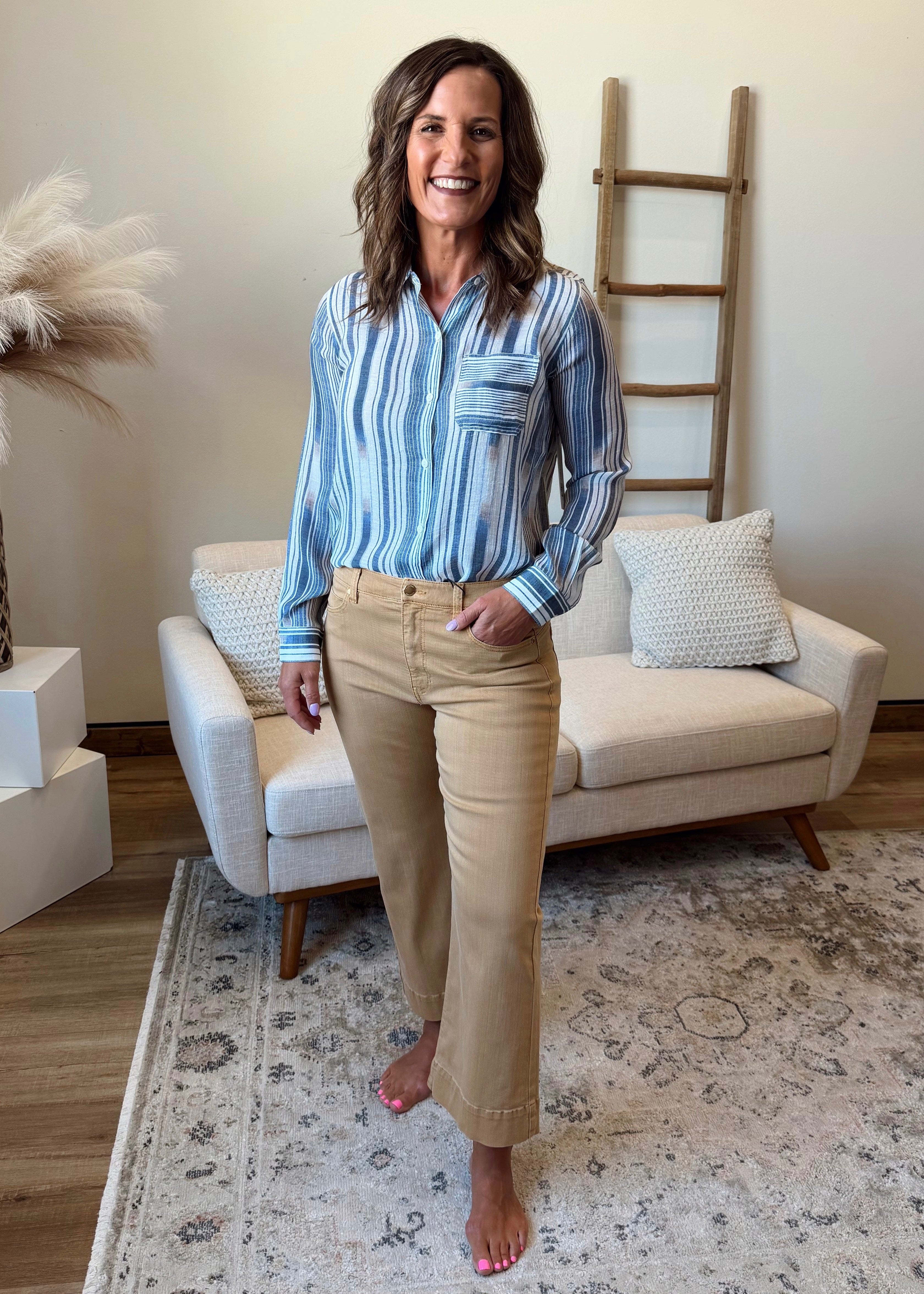 Woman standing in a living room wearing a blue striped shirt and beige pants.